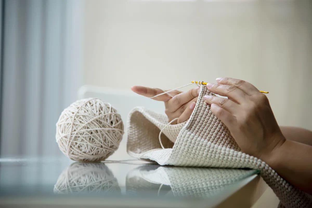 A pair of hands crocheting a cream panel with a yellow hook, a wound ball of the same yarn beside them.