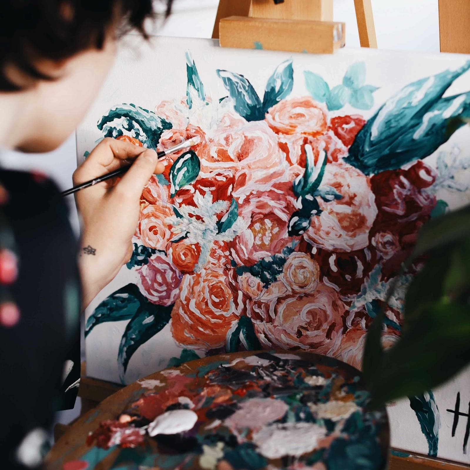 A painter at an easel, brush in hand, working into a canvas of coral and blush roses among deep teal leaves, a loaded palette in the foreground.