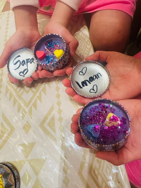Four handmade keepsakes cupped in children’s hands, two lettered with names and hearts and two filled with purple glitter and heart charms.