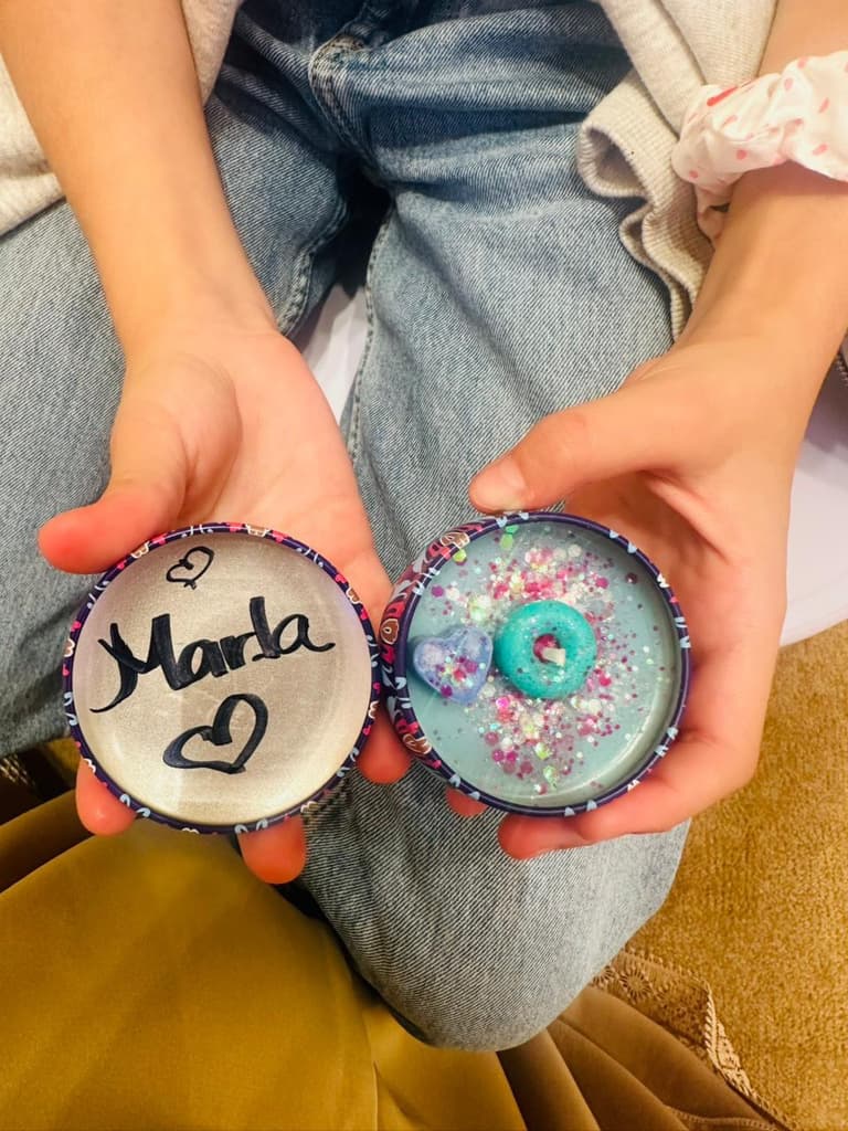 A child’s hands holding two handmade keepsakes: one lettered with a name and a heart, the other filled with glitter, sequins and a small teal ring.