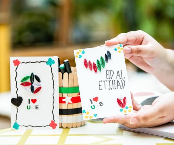 Two quilled cards for Eid Al Etihad reading “I love UAE”, beside a pen pot made from lolly sticks painted in the colours of the UAE flag.