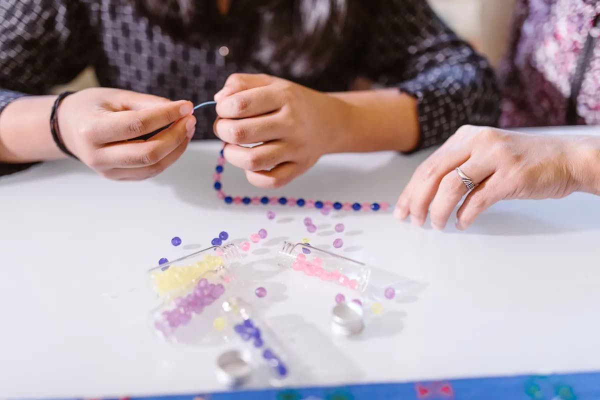 Two people at a white table threading a line of blue and red beads, loose beads and tubes in front of them.