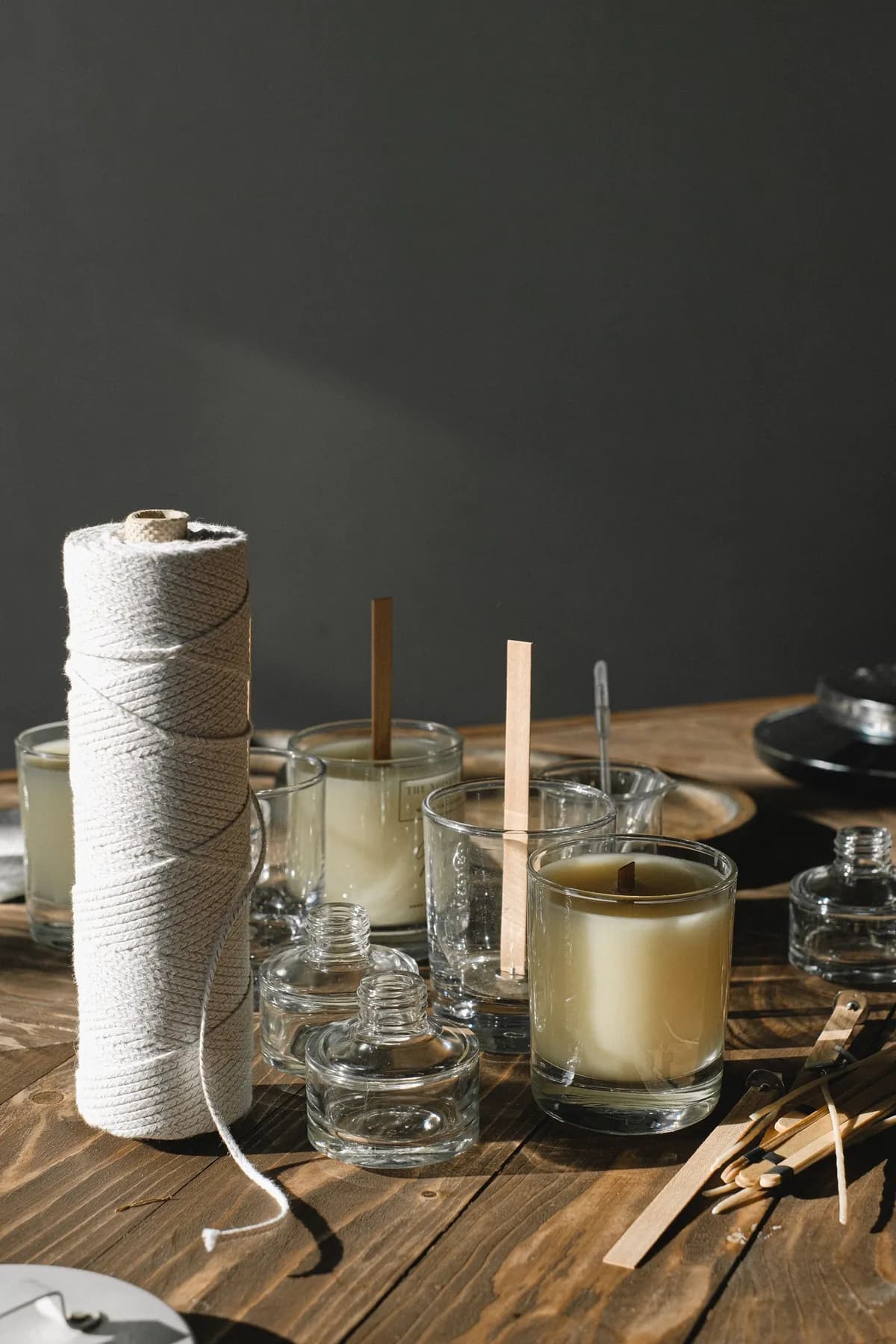 A wooden table set for pouring: filled candle glasses with their wicks held upright, empty jars and a spool of cream twine.