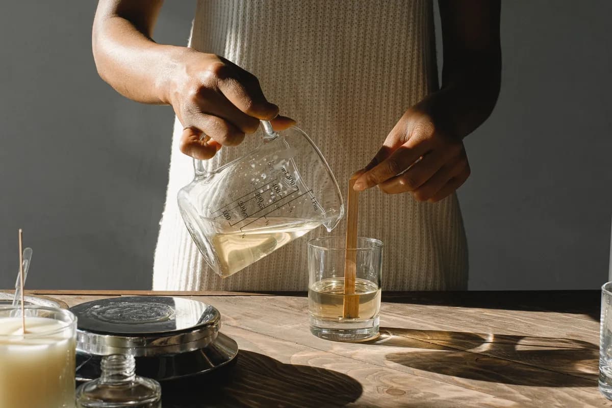 Wax poured from a glass jug into a tumbler while the other hand holds the wooden wick upright.