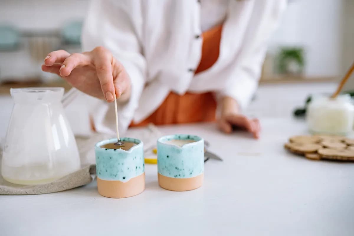 A hand setting a wick into one of two speckled turquoise and terracotta candle vessels on a white table.