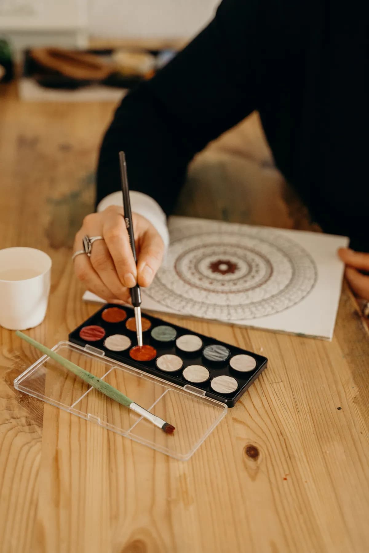 Someone at a wooden table drawing a mandala in fine concentric rings, a tray of watercolour pans beside them.