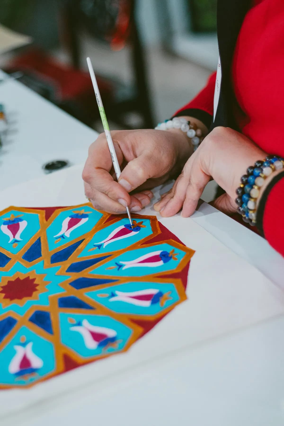 A hand painting a mandala of blue, pink and red petals radiating from a small sun at its centre.