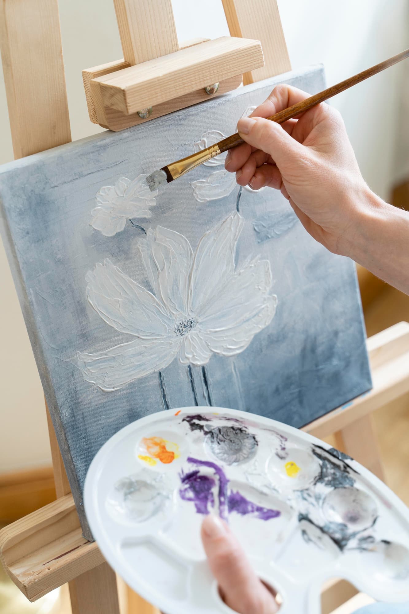 A hand drawing a brush across a small canvas on an easel, working a white bloom over a soft blue ground, a loaded palette below it.