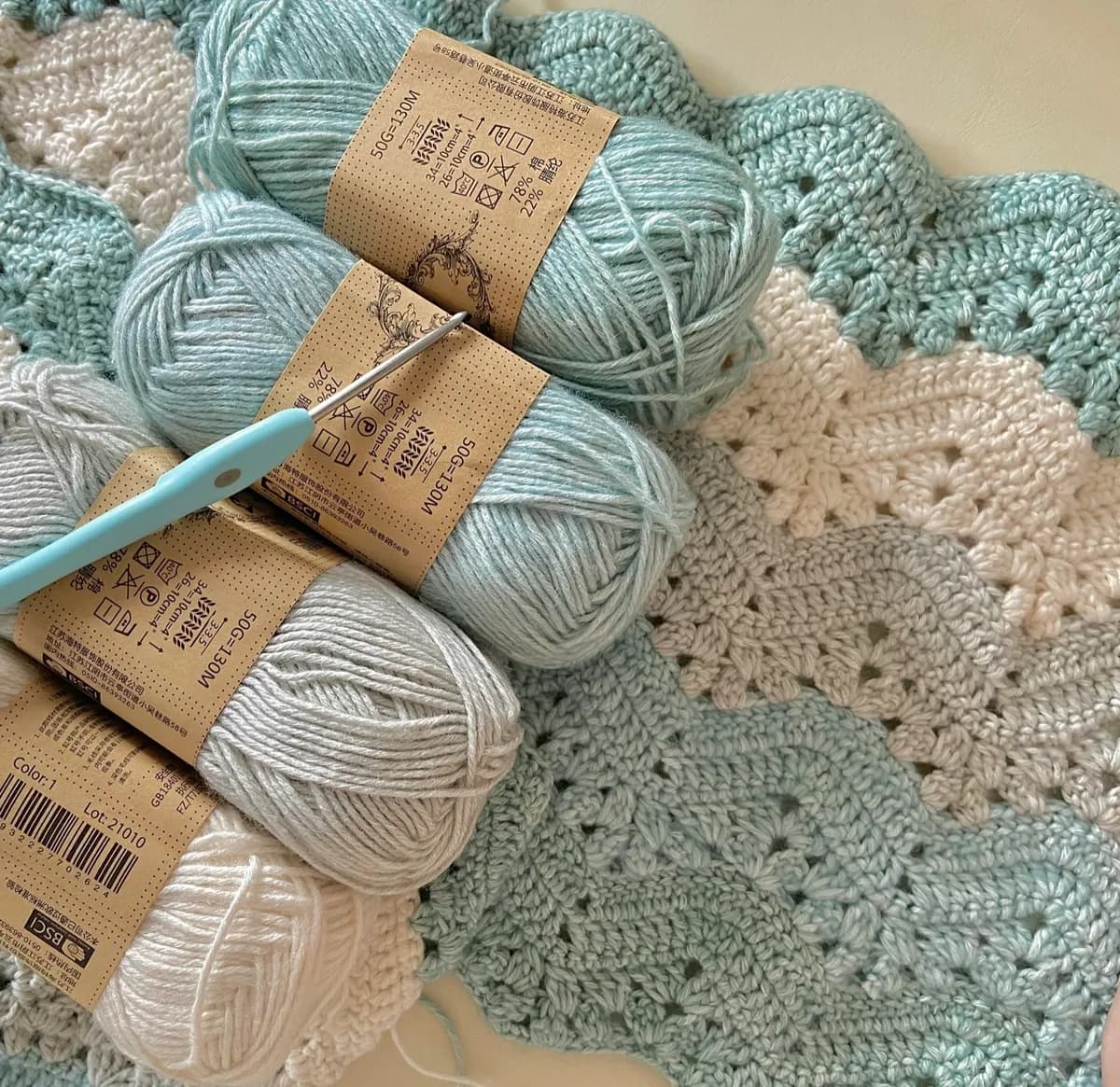 Balls of pale blue and cream yarn with a crochet hook resting on a finished blanket of shell stitch.
