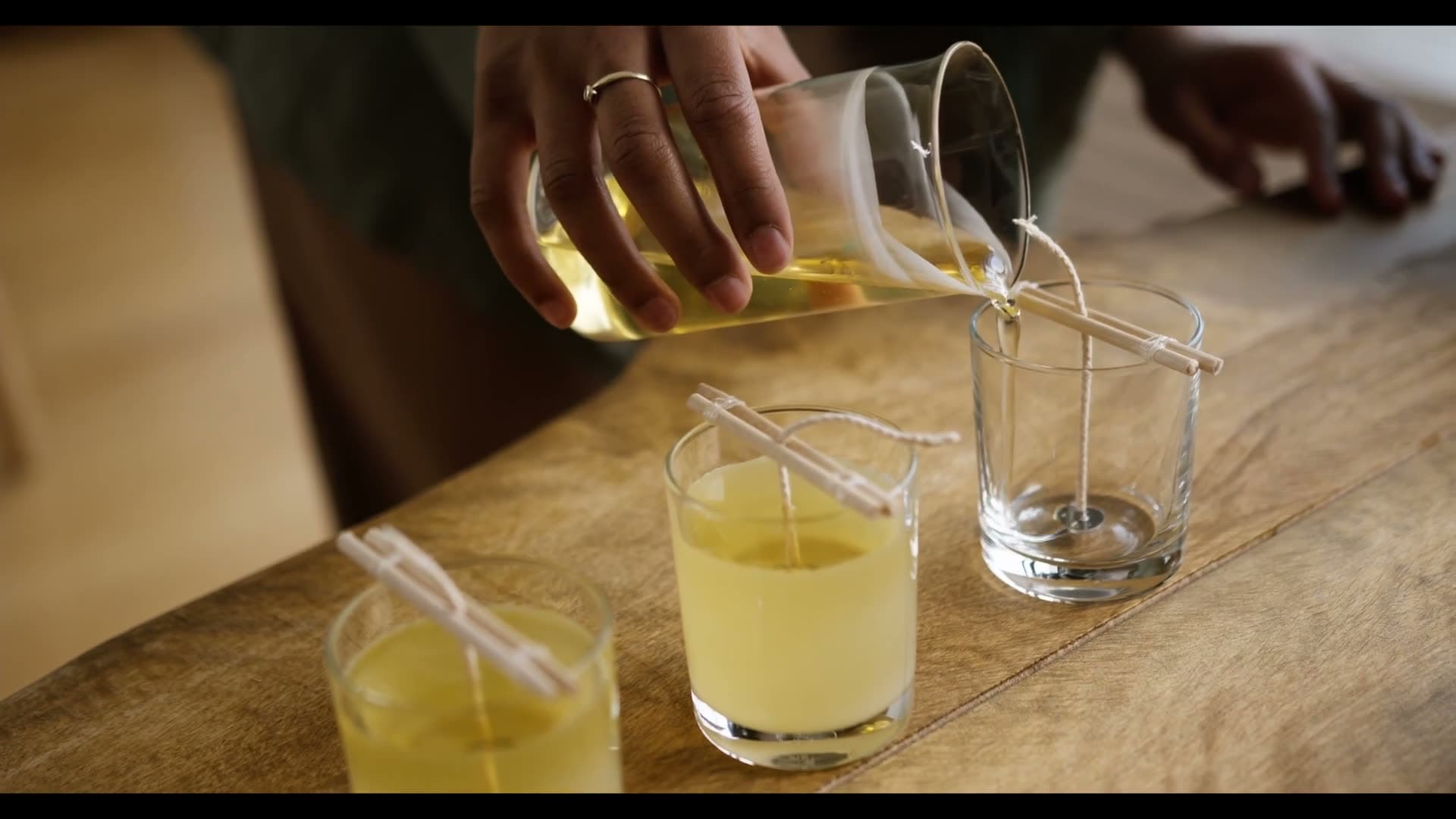 Wax being poured from a jug into a row of glass candle jars on a workshop table.