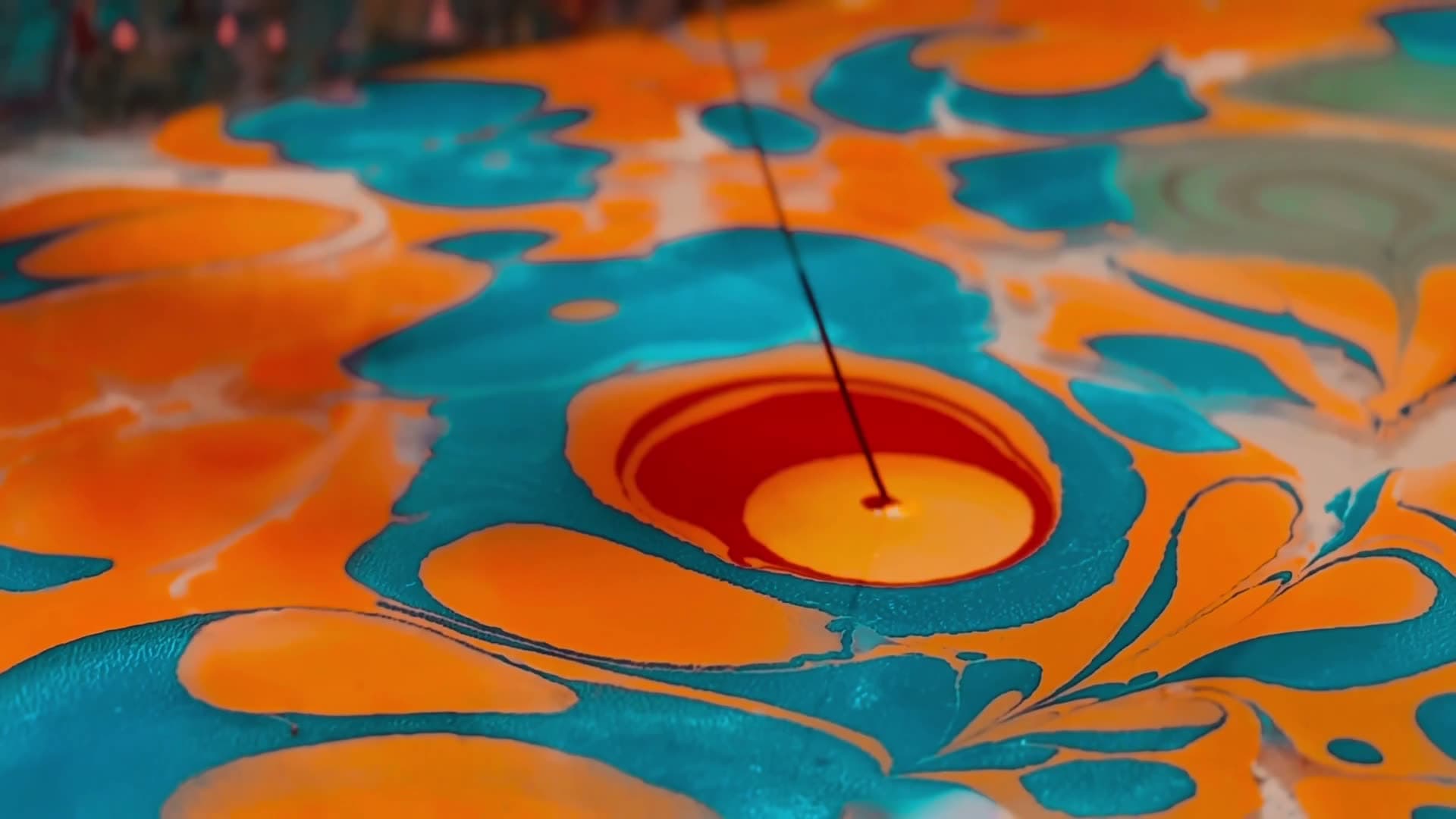 A close view of a marbling bath: a fine needle drawn down through floating orange, teal and red inks, pulling them into feathered swirls.