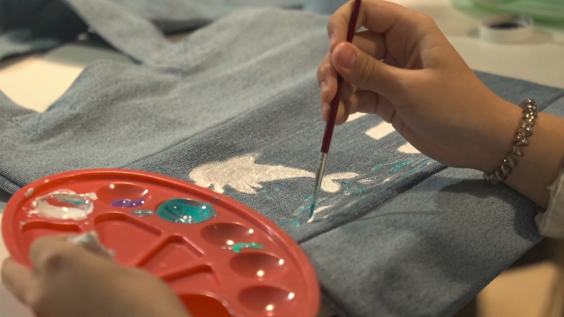 A hand painting a silver motif onto denim with a fine brush, a red palette of mixed colour beside it.