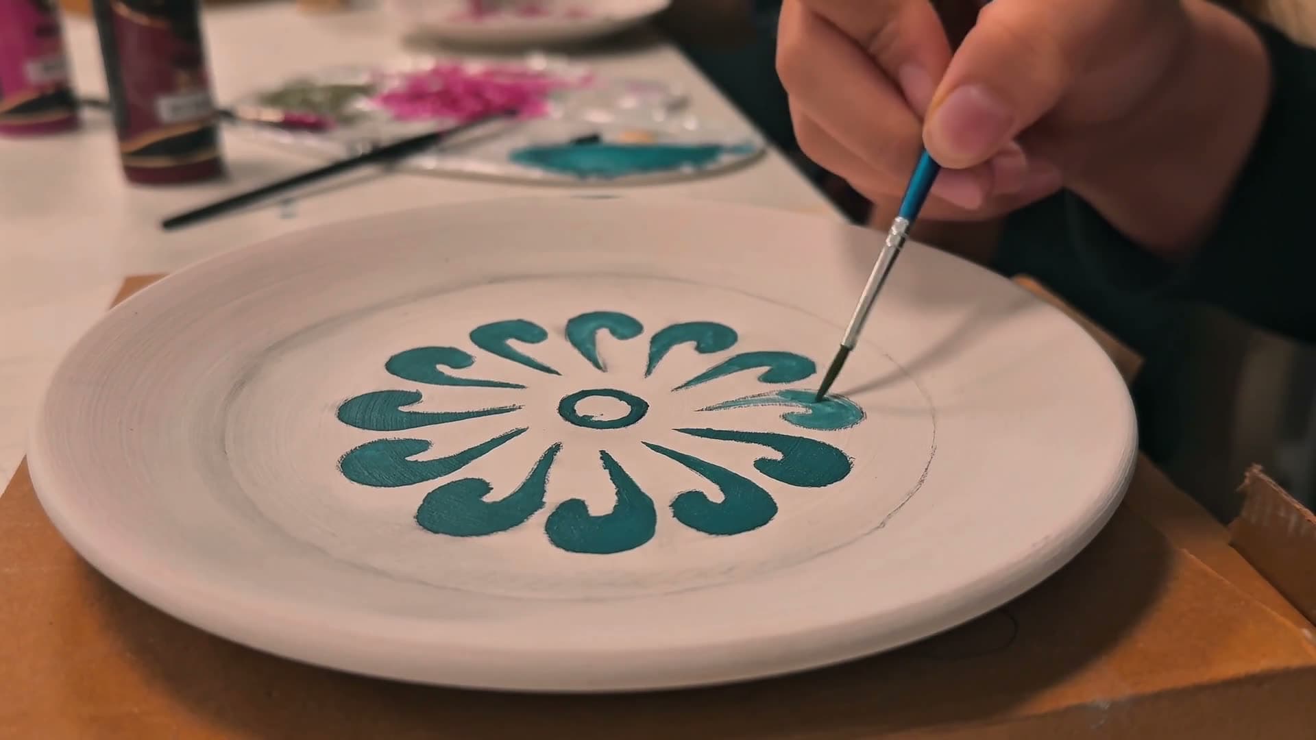 A hand painting a teal flower motif onto a pale ceramic plate.