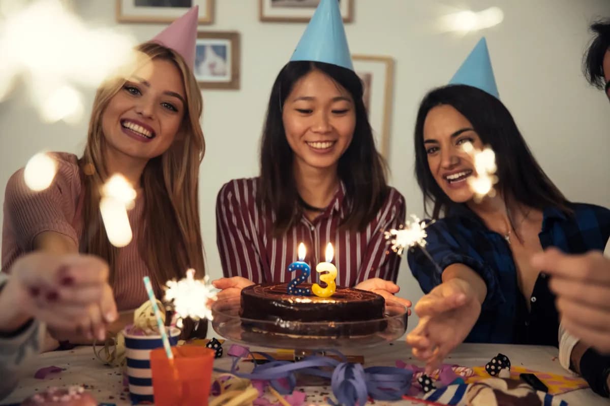 Three friends in paper party hats leaning in around a chocolate birthday cake with a lit number candle, sparklers burning in their hands.