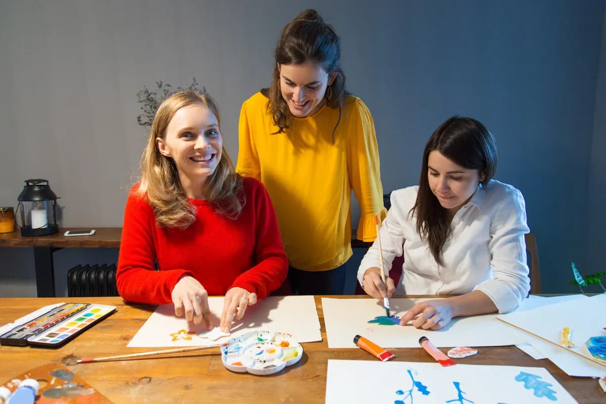 Three colleagues around a wooden table painting on paper with watercolours, one standing and leaning in over the other two.