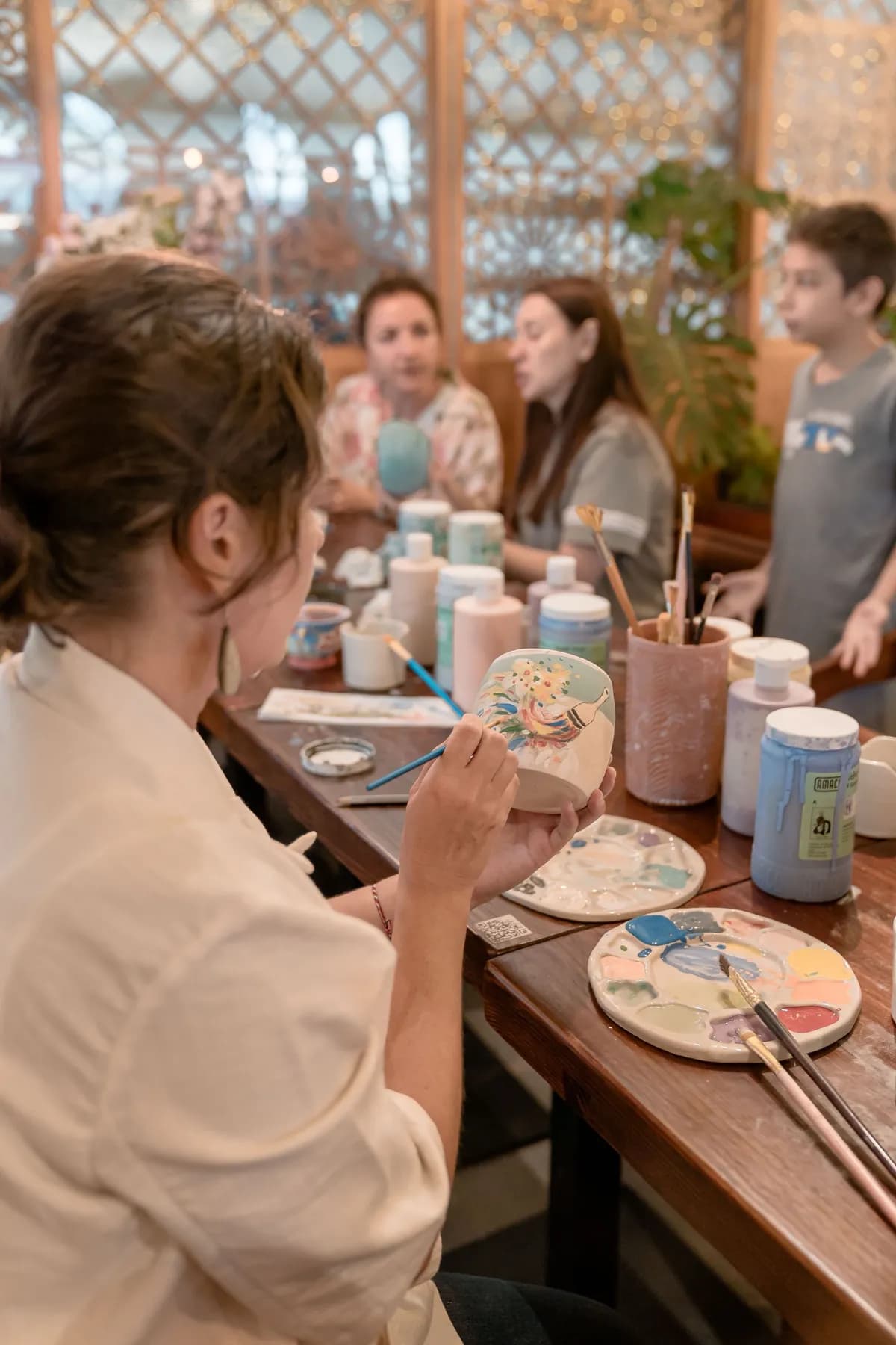 A woman painting a pattern onto a ceramic mug at a long table of glaze bottles and brush pots, with other people working and talking behind her.