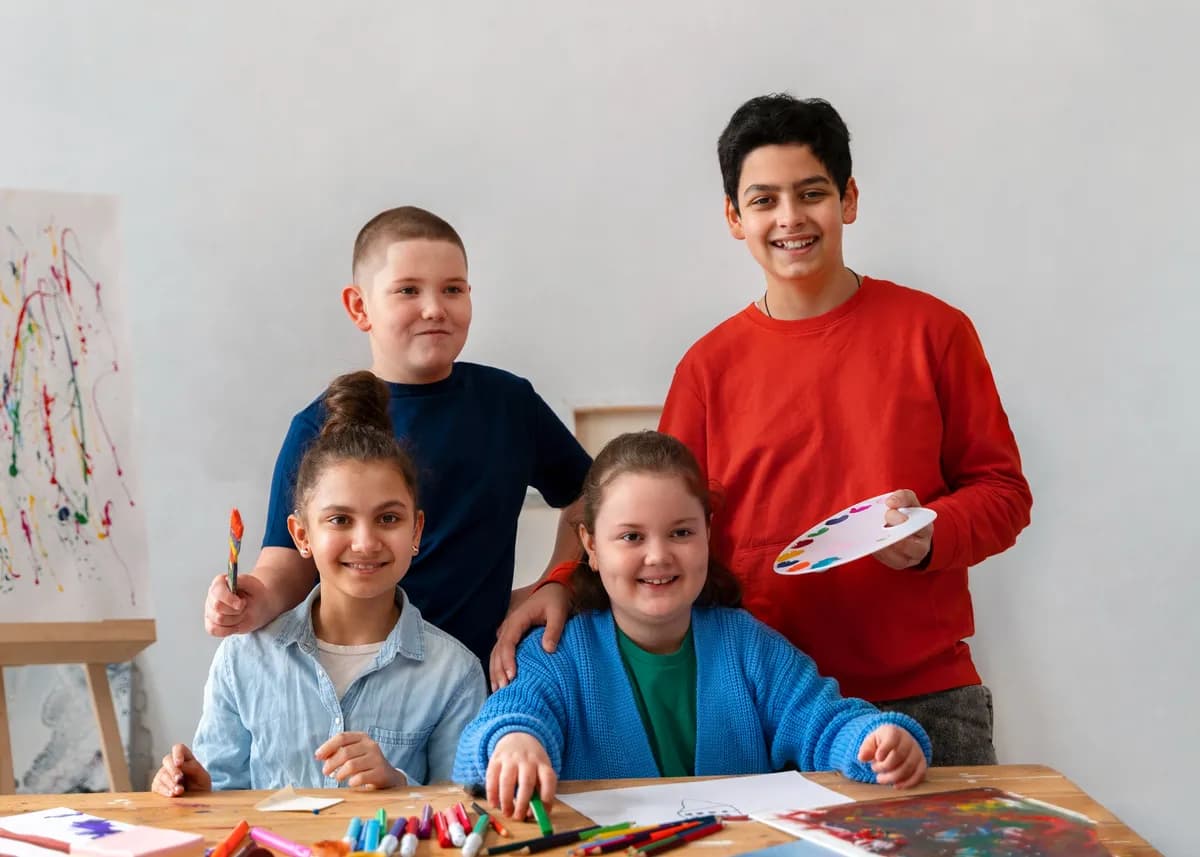 Four school-age children at a table spread with felt tips and paintings, one holding up a loaded paint palette and another a brush.