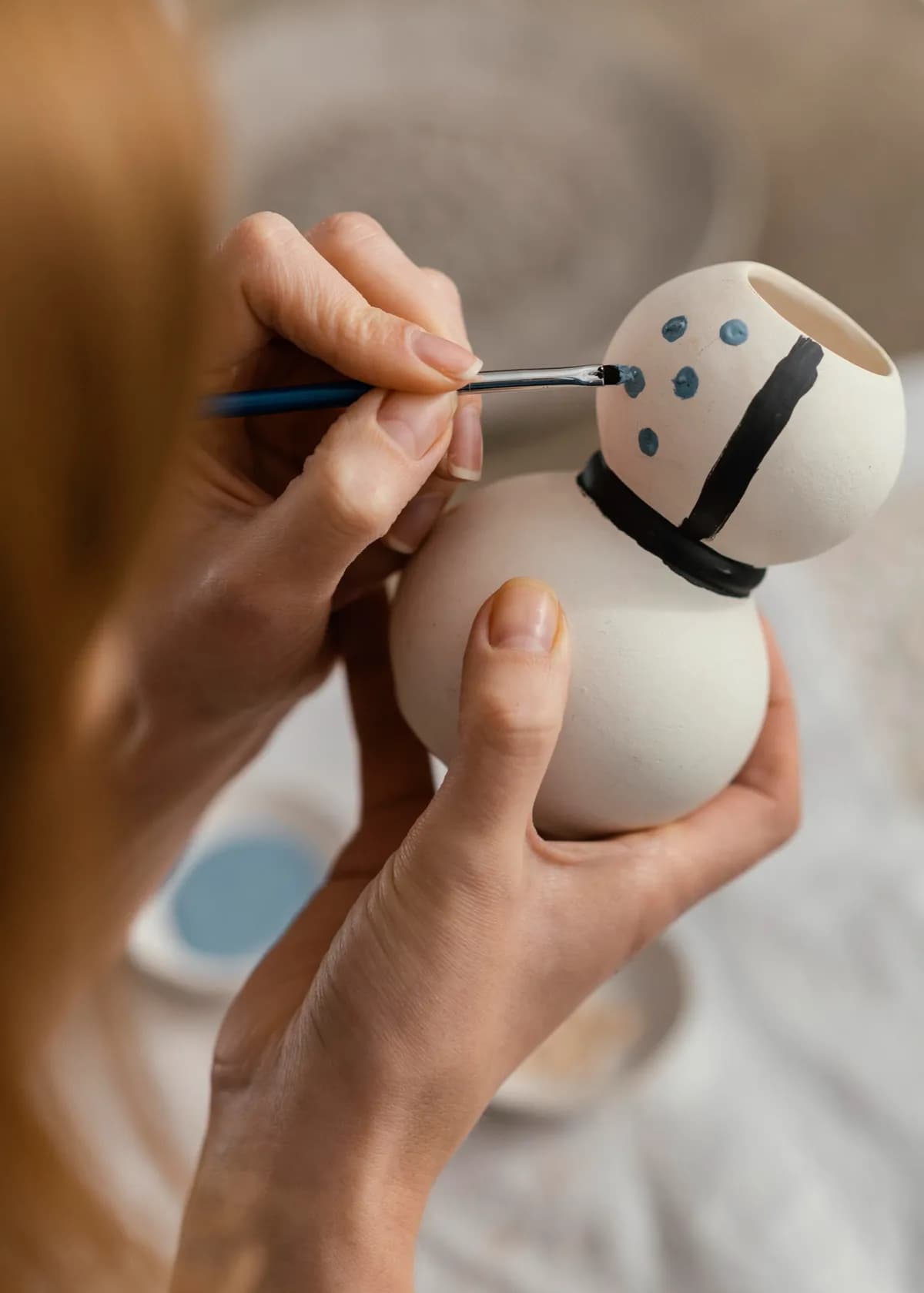 Hands holding a small unglazed pot while a fine brush lays a band of blue dots around its neck.