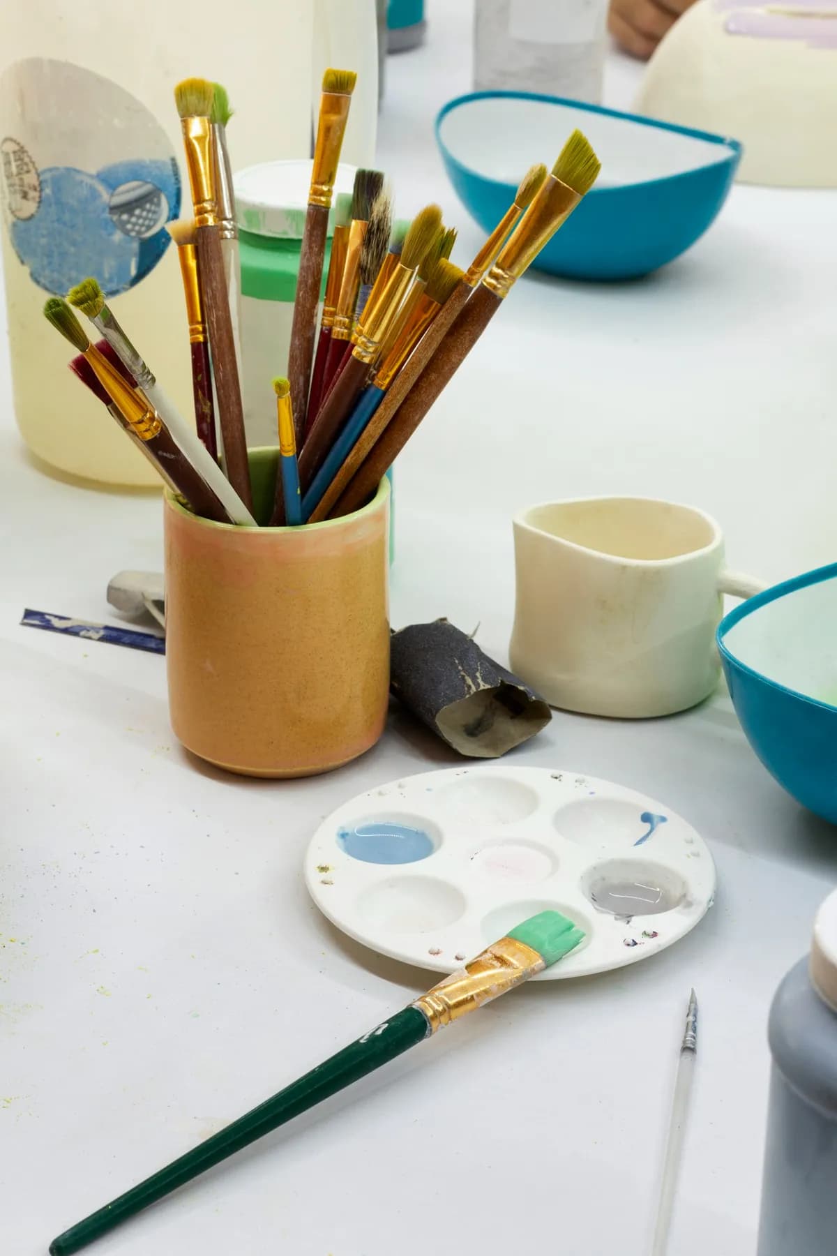 A jar of brushes, a palette of mixed blues and greys, and unpainted cups and bowls set out on a white table.