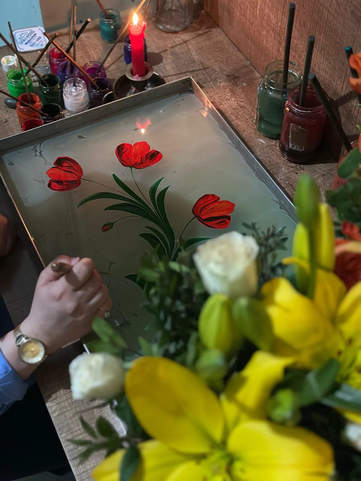 A hand painting red poppies and green stems onto a glass panel laid flat, jars of colour around it.