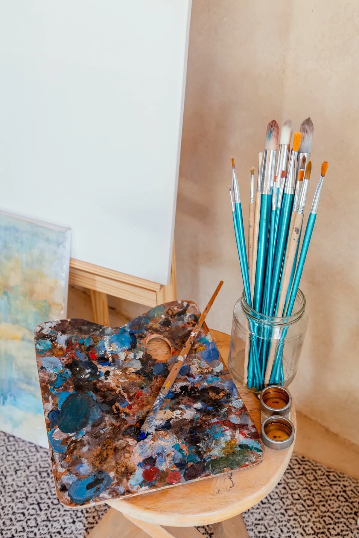 Brushes standing in a jar beside a paint-smeared wooden palette on a small round table, an easel behind.