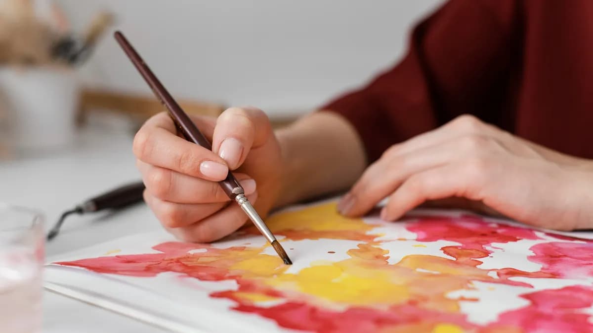 A hand laying loose red and yellow blooms of colour across a sheet of paper, the other hand steadying the page.