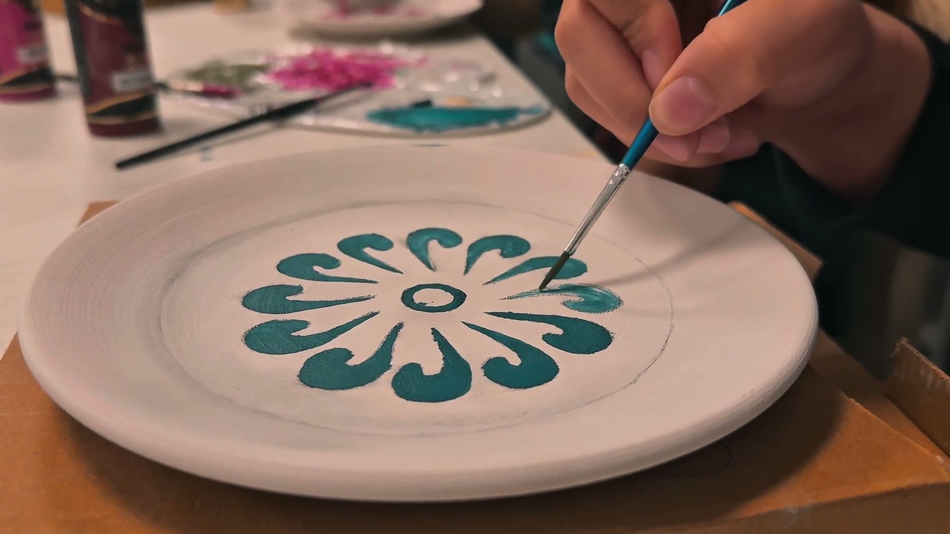 A pair of hands painting a ceramic plate at a table, a brush in one of them.
