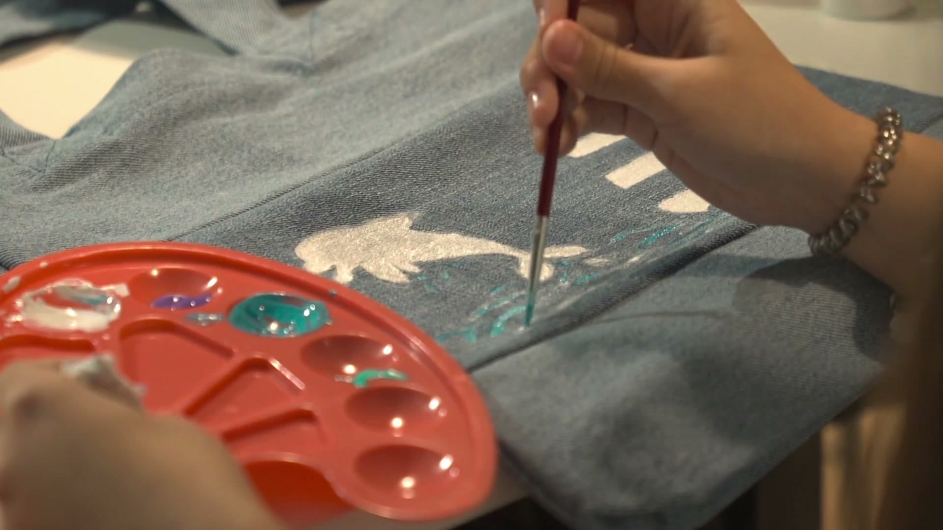 Two pairs of hands at one table: one holds a red paint palette, the other paints a silver and teal design onto a pale denim tote bag.