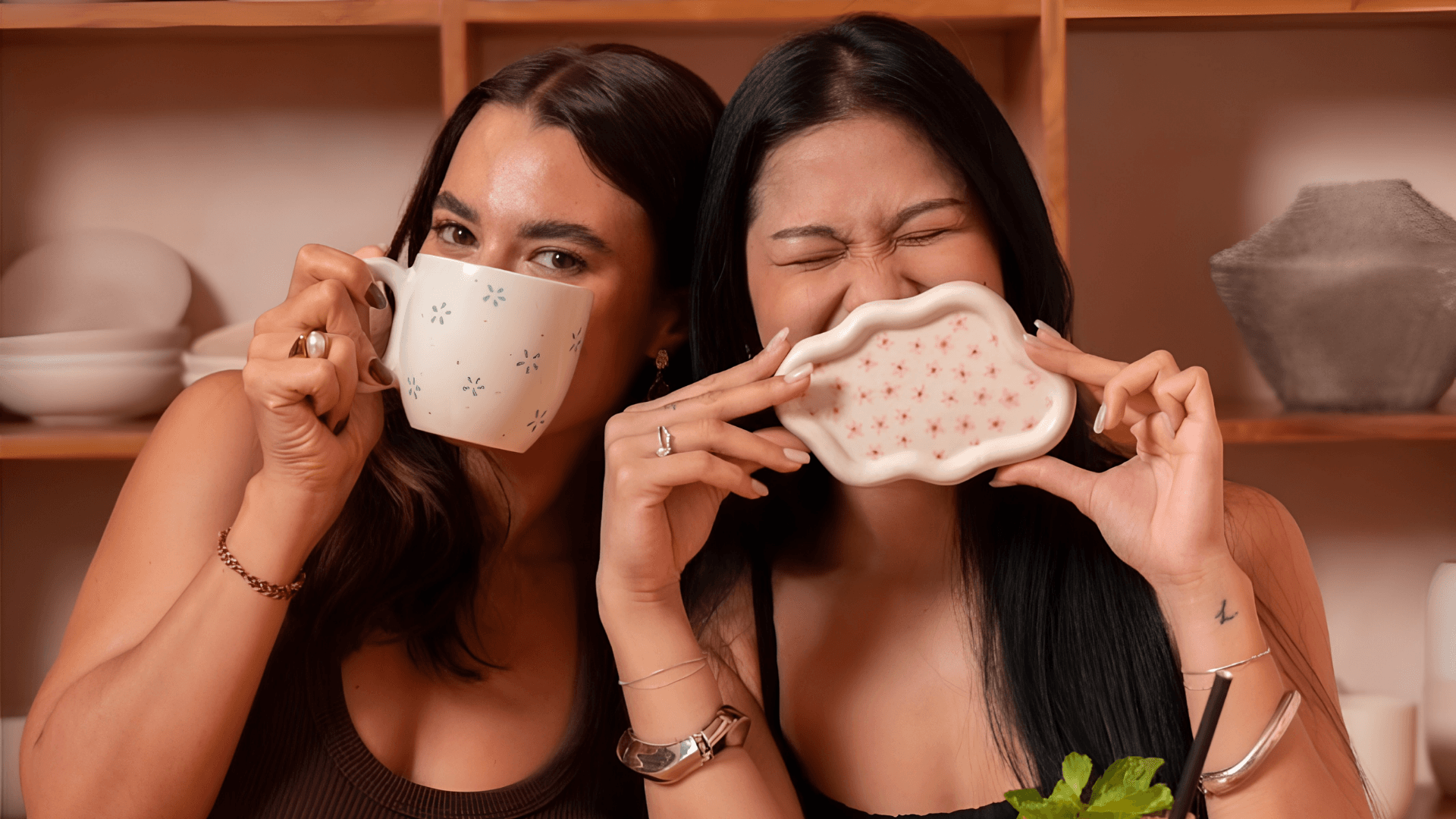 Two women laughing behind the ceramics they have painted, one holding up a mug dotted with small blue flowers, the other a scallop-edged tray patterned with little pink blooms, with shelves of plain crockery behind them.