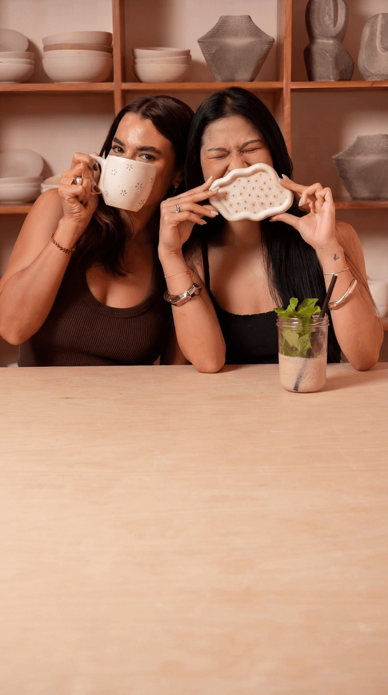 Two women laughing behind the ceramics they have painted, one holding up a mug dotted with small blue flowers, the other a scallop-edged tray patterned with little pink blooms, at a wooden table with shelves of plain crockery behind them.